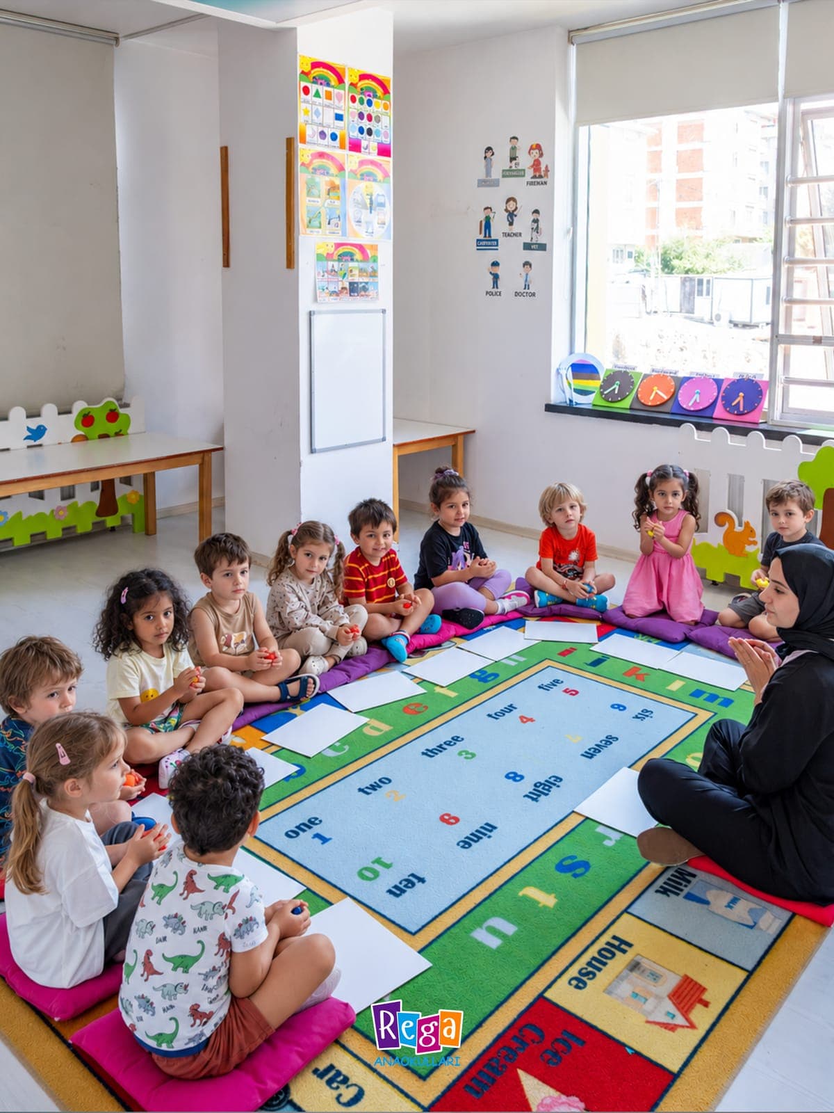 İngilizce sayı halısının çevresinde öğretmenleriyle oturan çocuklar